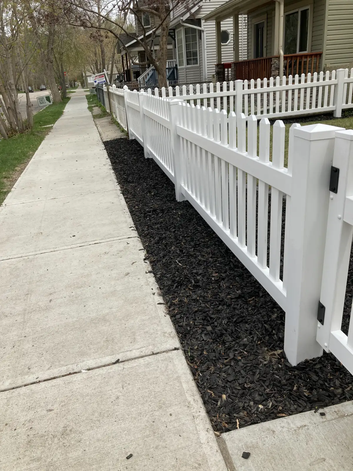 A white vinyl picket fence with scalloped tops and a gate post, built along a Sunnyside sidewalk in Calgary