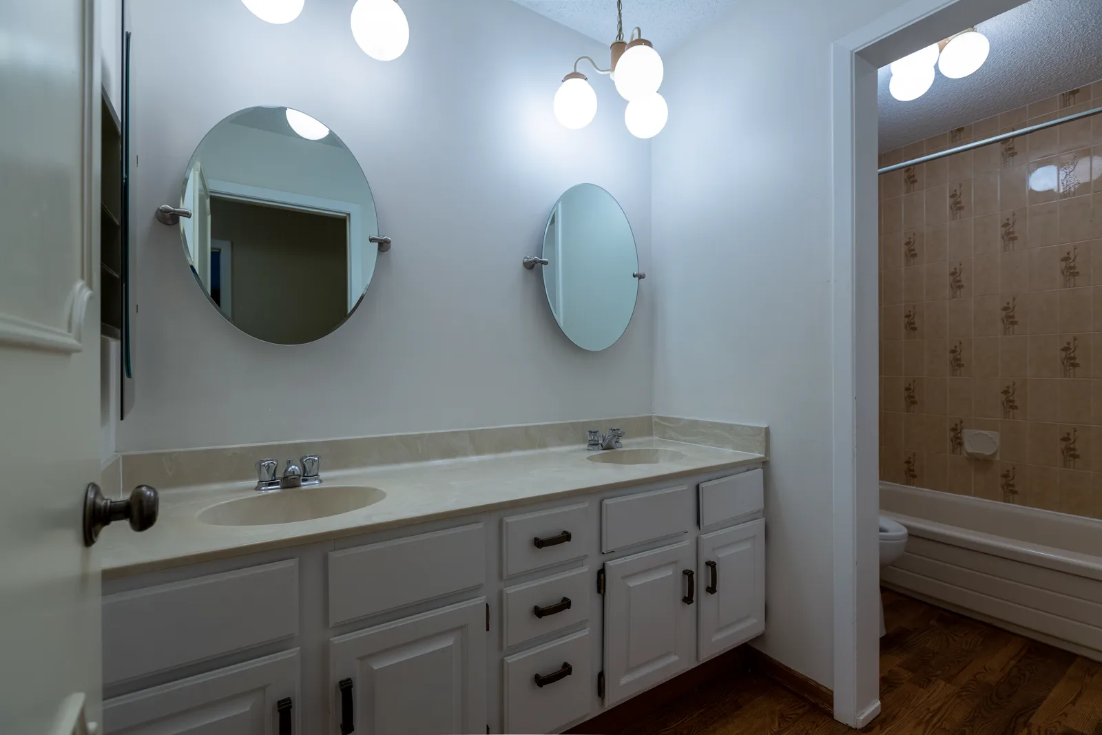 Rideau bathroom renovation in Calgary, before — a cultured-marble double vanity under a cluster of white glass globe lights with a beige floral tub tile alcove
